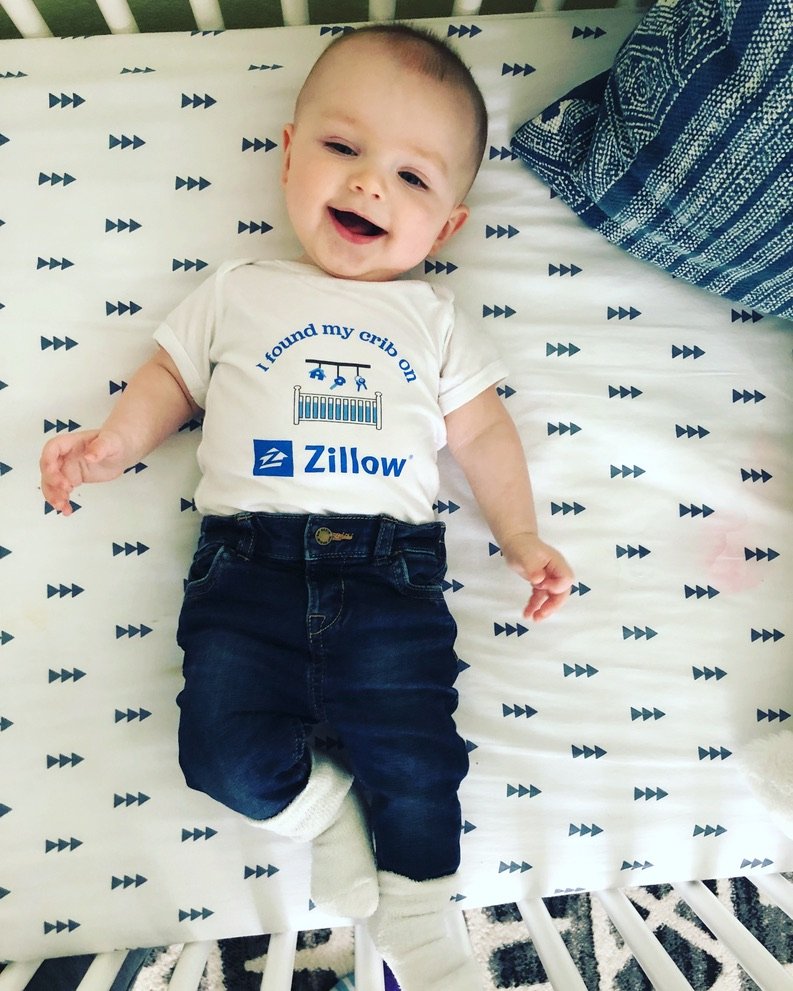 A smiling baby in a Zillow onesie that reads I found my crib on Zillow